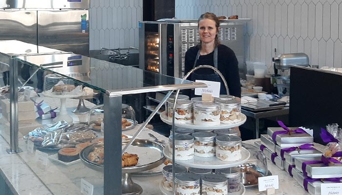 A woman stands behind a café dessert counter with cakes, pastries, and treats in jars on display.